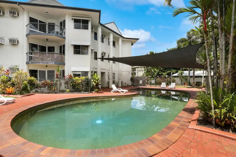 Piscine rectangulaire entourée d'une terrasse carrelée et de végétation tropicale devant un bâtiment blanc.
