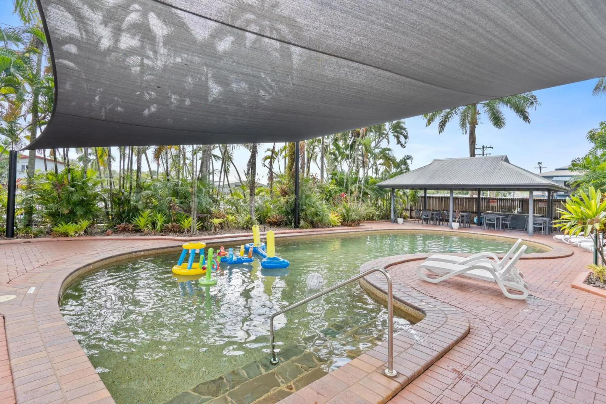 Piscine rectangulaire avec jeux flottants et transats sous un auvent ombragé.
