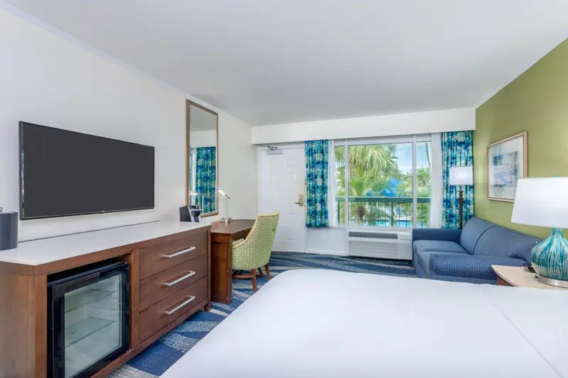 Une chambre lumineuse avec un lit, un bureau et un canapé face à une baie vitrée.