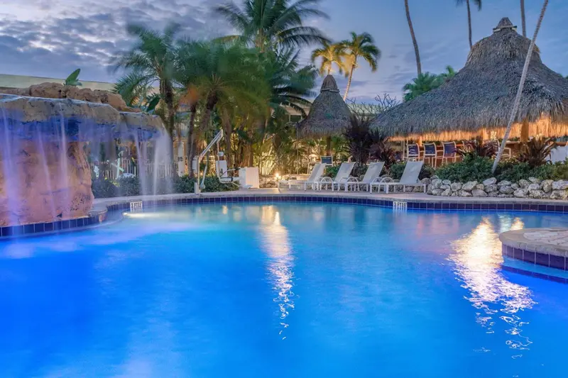 Une piscine illuminée au crépuscule, avec des chaises longues et des toits de chaume en arrière-plan.