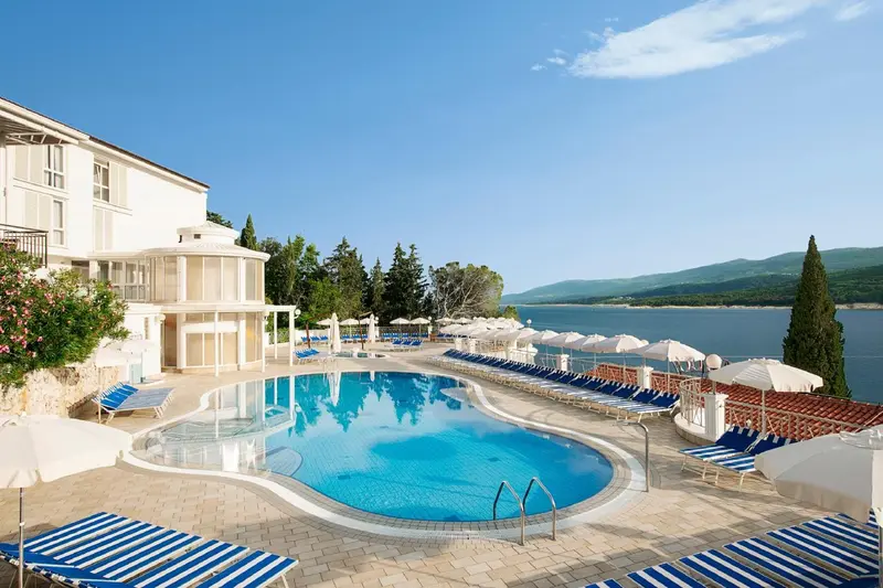 Piscine d'hôtel avec transats et parasols, vue sur la mer et les montagnes.