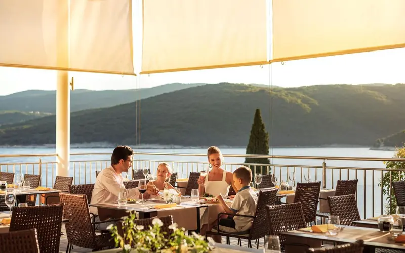Une famille prend son repas sur une terrasse ensoleillée avec vue sur la mer et les montagnes.