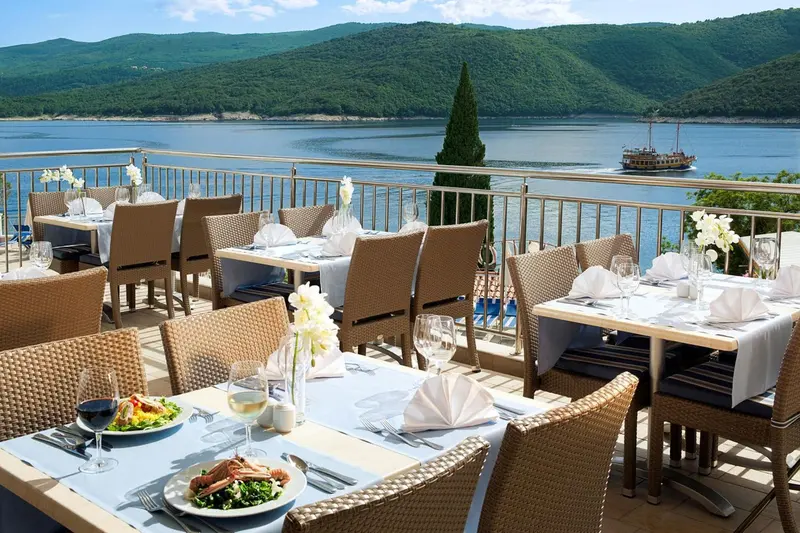 Une terrasse avec des tables dressées pour un repas en plein air offre une vue sur la mer et les collines.