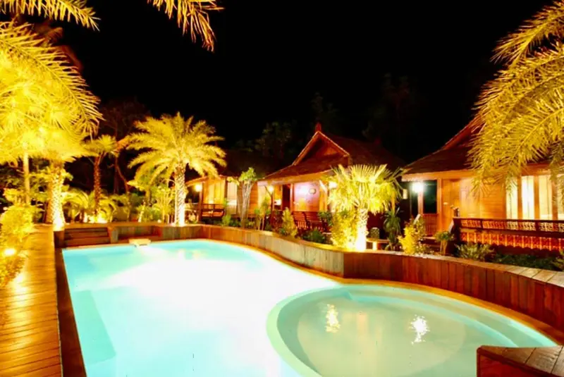 Une piscine illuminée au premier plan, avec des bungalows éclairés en arrière-plan.