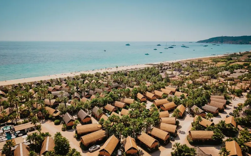 Vue aérienne d'une plage bordée de bungalows aux toits de chaume et de végétation luxuriante.