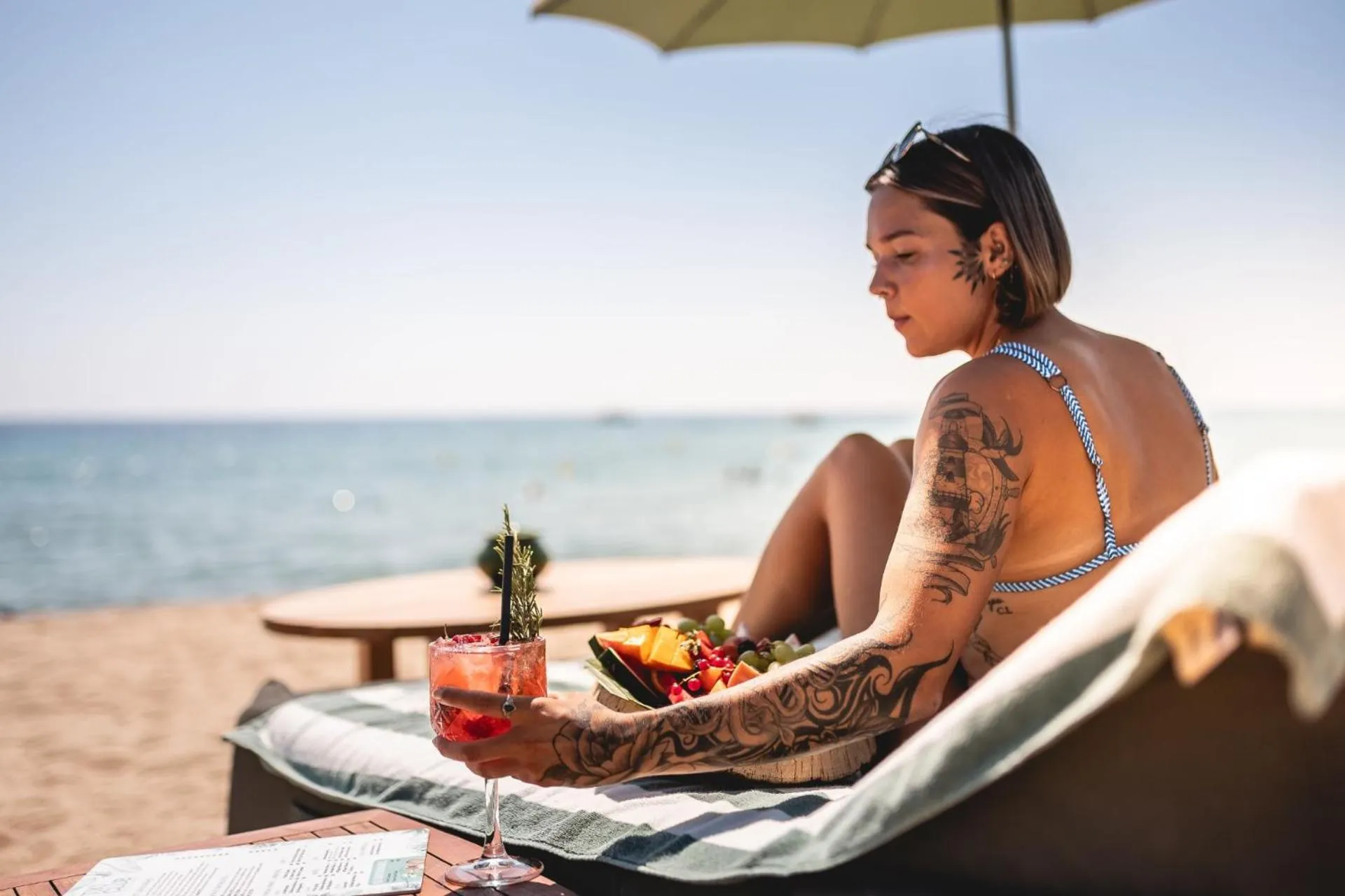 Une personne se détend sur une chaise longue avec un cocktail et des fruits sur une plage.