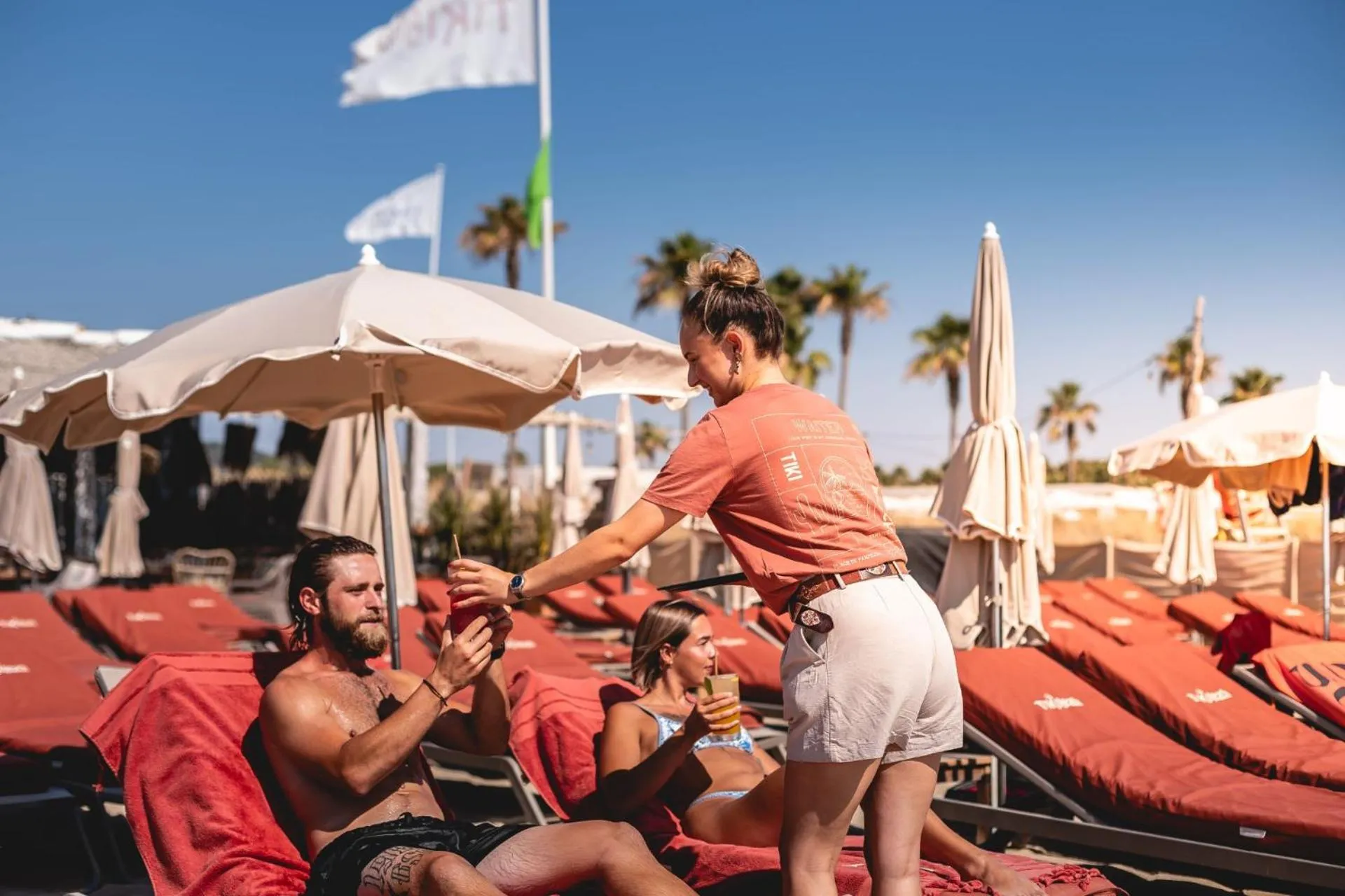 Une serveuse apporte des boissons à des clients allongés sur des transats rouges près d'une piscine.