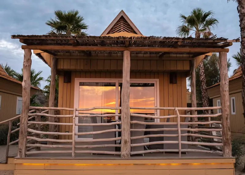 Façade d'un bungalow en bois avec une terrasse et un coucher de soleil en arrière-plan.