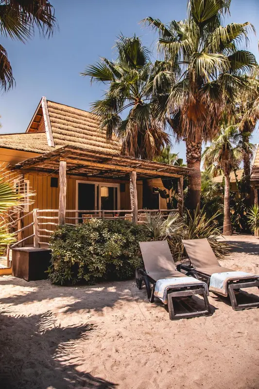 Deux transats invitent à la détente sur une plage de sable fin devant un bungalow au toit de chaume.