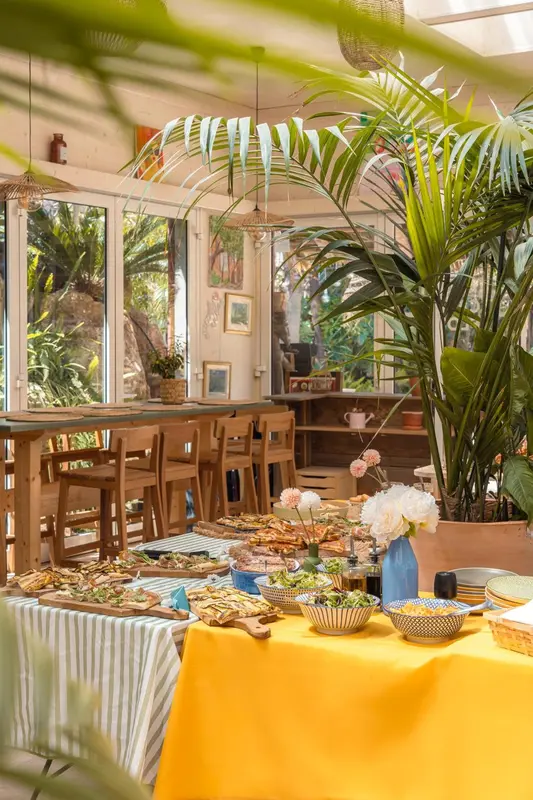 Une table garnie de mets variés est présentée dans une salle lumineuse décorée de plantes.