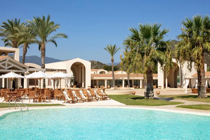 Vue d'une piscine devant un bâtiment blanc avec des palmiers et des transats.