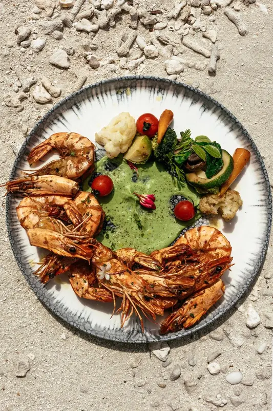 Assiette de crevettes grillées et de légumes servis avec une sauce verte, vue d'en haut sur du sable.