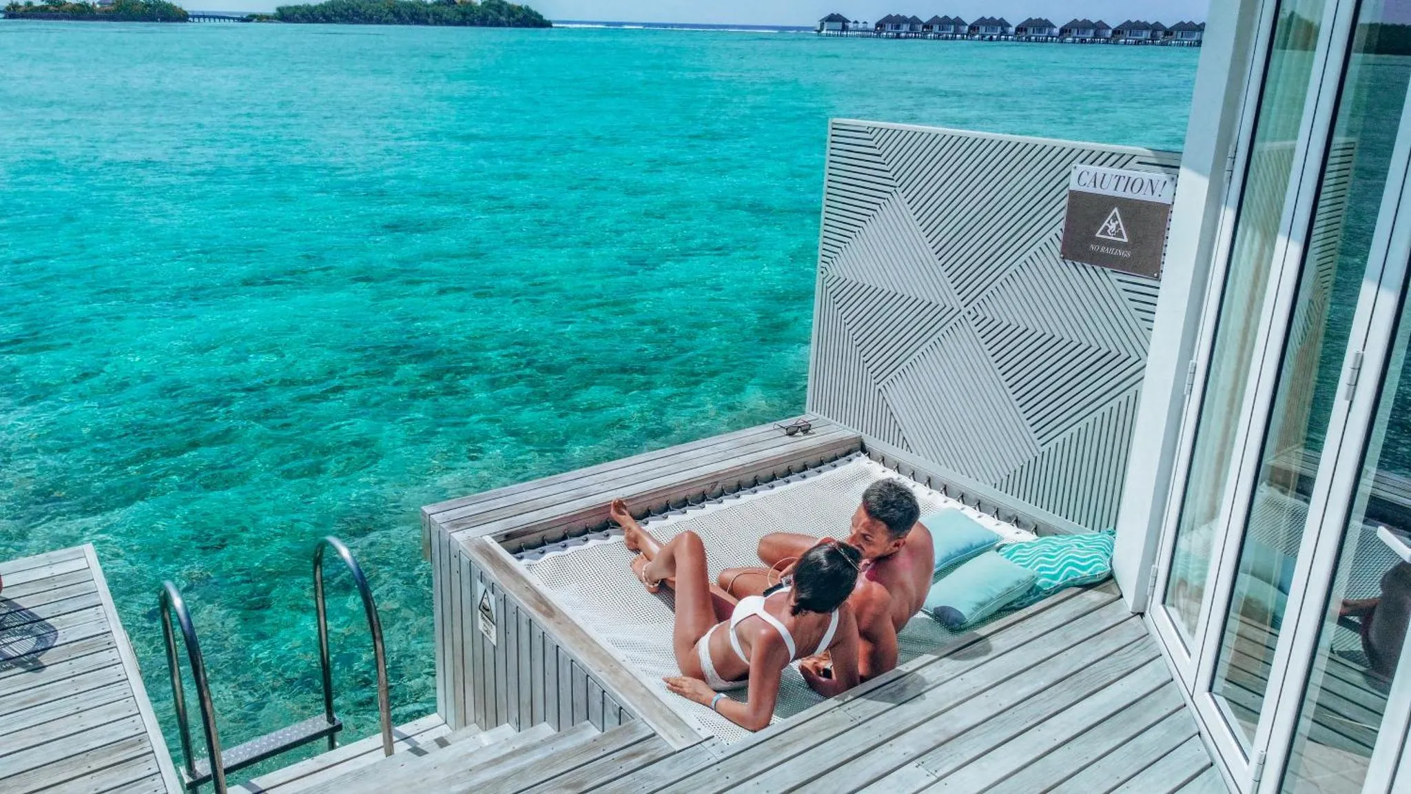 Deux personnes se détendent sur une terrasse en bois au-dessus de l'eau turquoise.