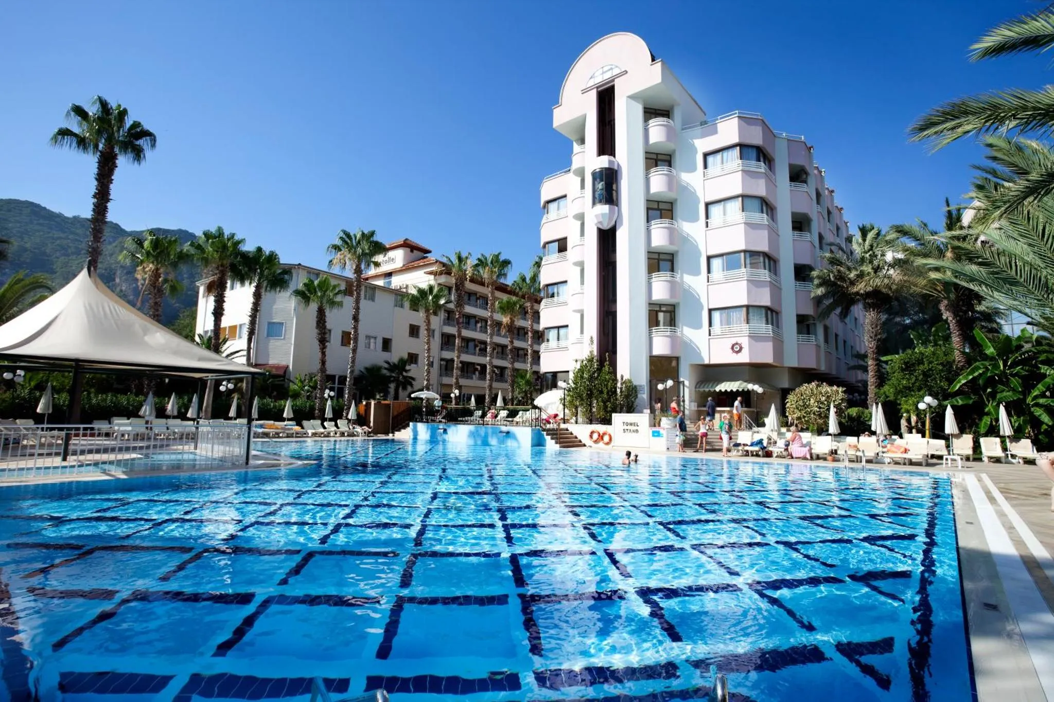 Vue d'une piscine d'hôtel avec des transats et des palmiers sous un ciel bleu.