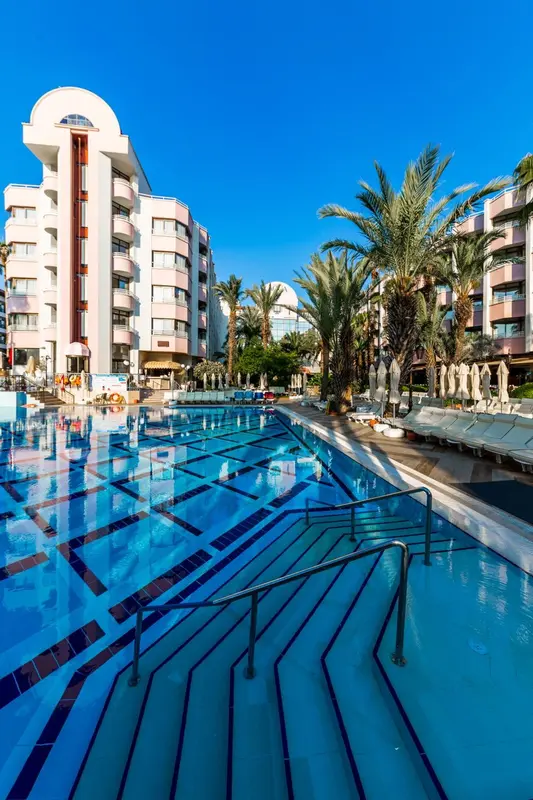 Une piscine rectangulaire turquoise avec des escaliers intégrés, bordée de transats et de palmiers, devant un bâtiment blanc et rose.
