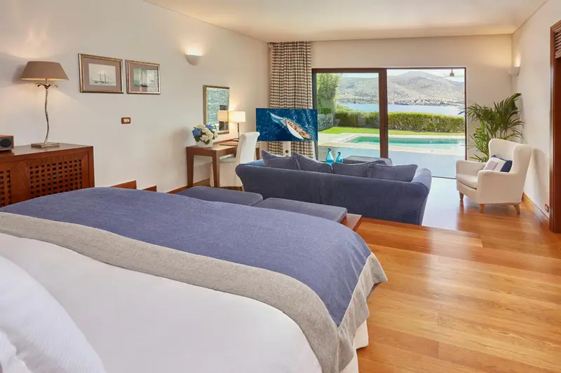 Une chambre lumineuse avec un grand lit et un espace salon donnant sur une piscine et la mer.