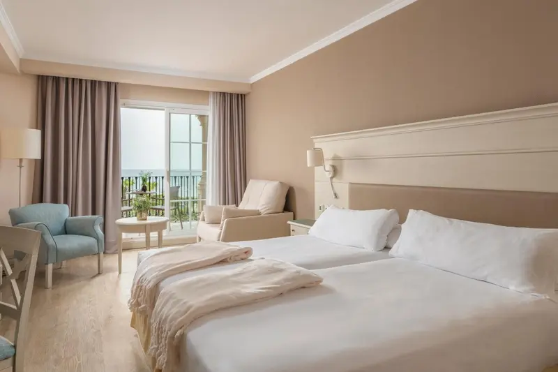 Chambre lumineuse avec un lit double et un coin salon donnant sur une vue extérieure.