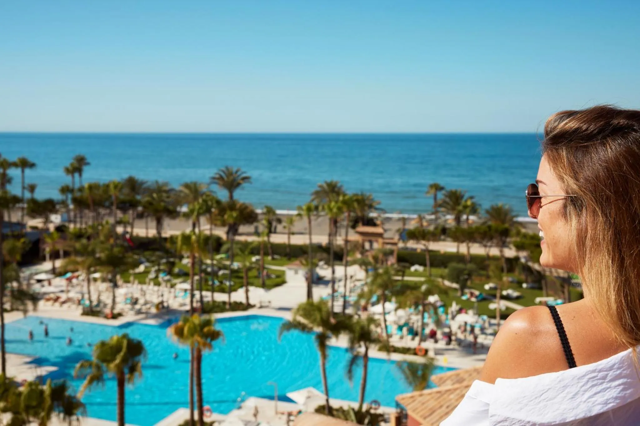 Une femme admire la vue sur la mer et la piscine depuis un balcon.