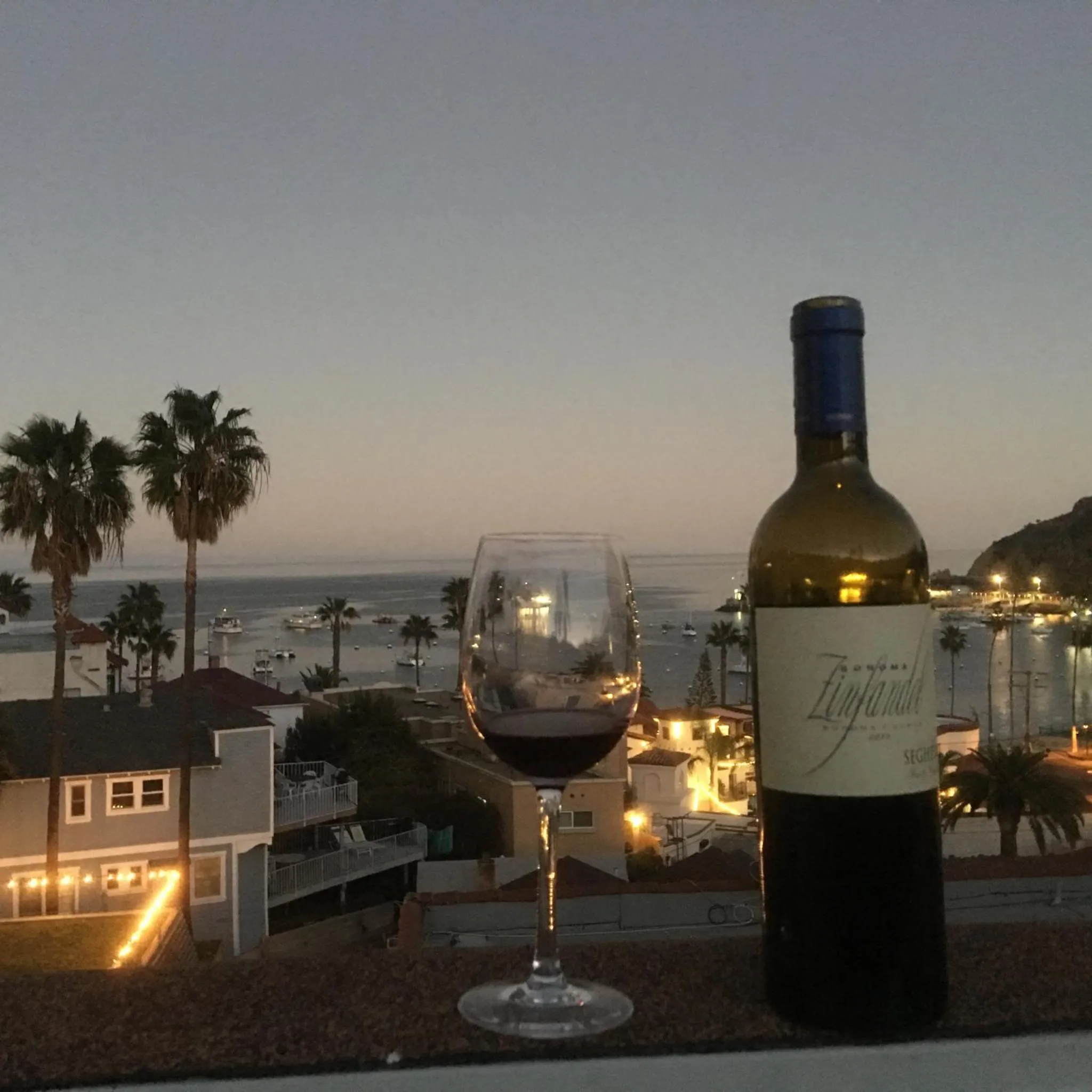 Une bouteille de vin rouge et un verre sur une terrasse avec vue sur la mer au coucher du soleil.