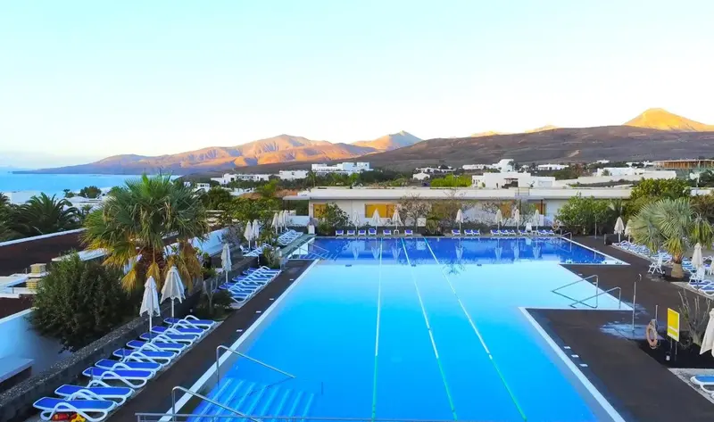 Vue aérienne d'une piscine rectangulaire bordée de transats, avec des montagnes en arrière-plan.