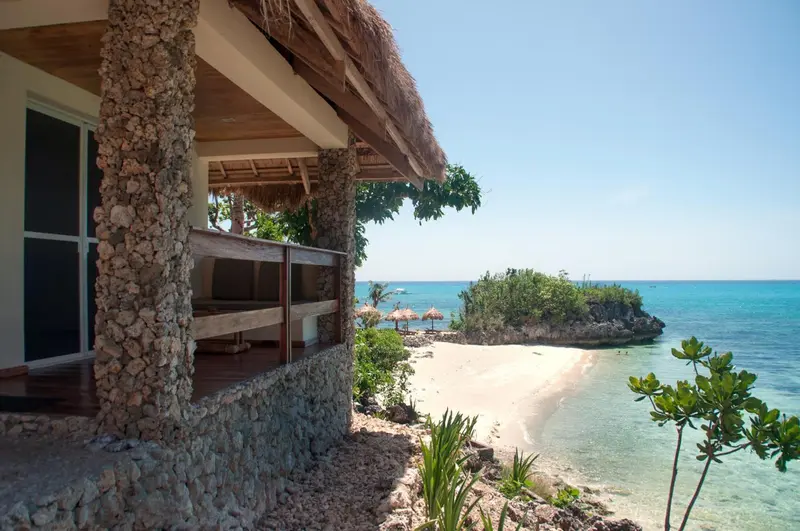 Terrasse en bois devant un bâtiment aux murs de pierre, surplombant une plage de sable blanc et une mer turquoise.