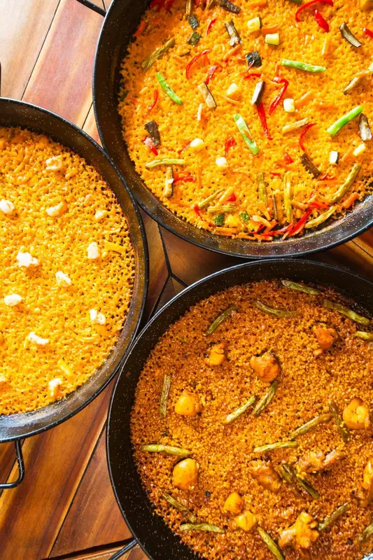 Trois grandes poêles de paella colorée sont présentées sur une table en bois.