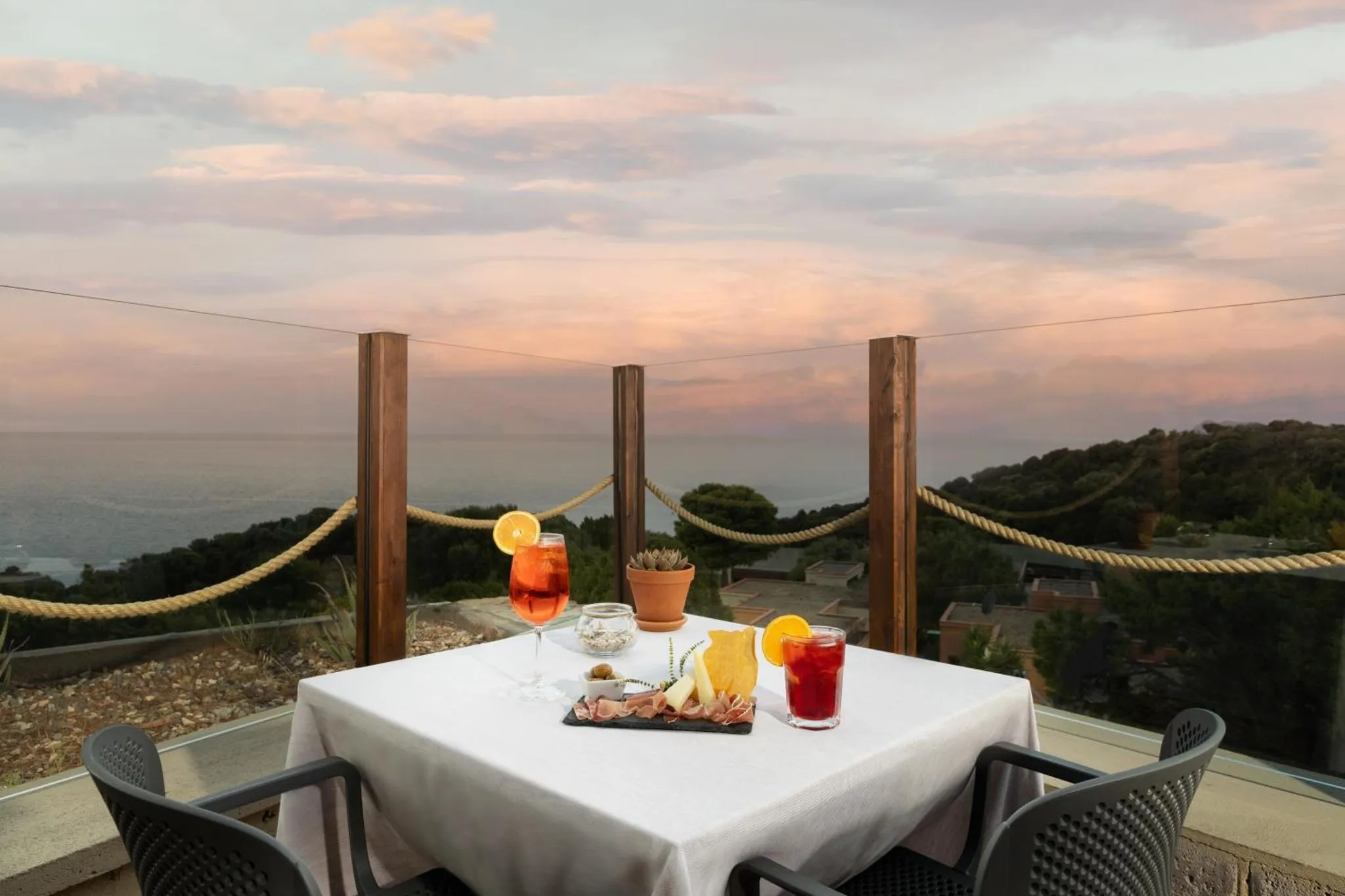 Table dressée avec des boissons et des amuse-gueules sur une terrasse avec vue sur la mer au coucher du soleil.