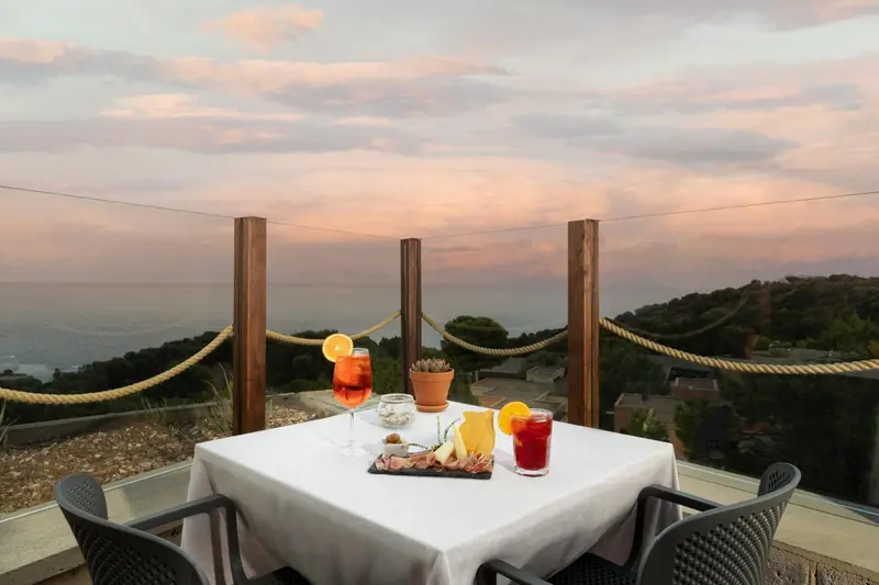 Table dressée avec des boissons et des amuse-gueules sur une terrasse avec vue sur la mer au coucher du soleil.