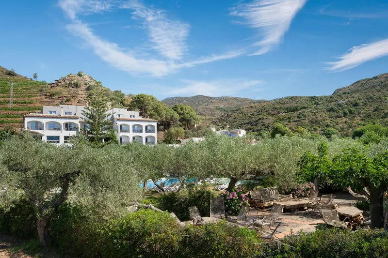 Vue d'un bâtiment blanc entouré de végétation et d'une piscine en contrebas.