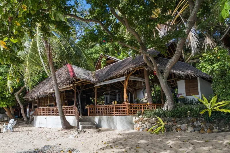 Bâtiment à toit de chaume avec terrasse en bois donnant sur une plage de sable.