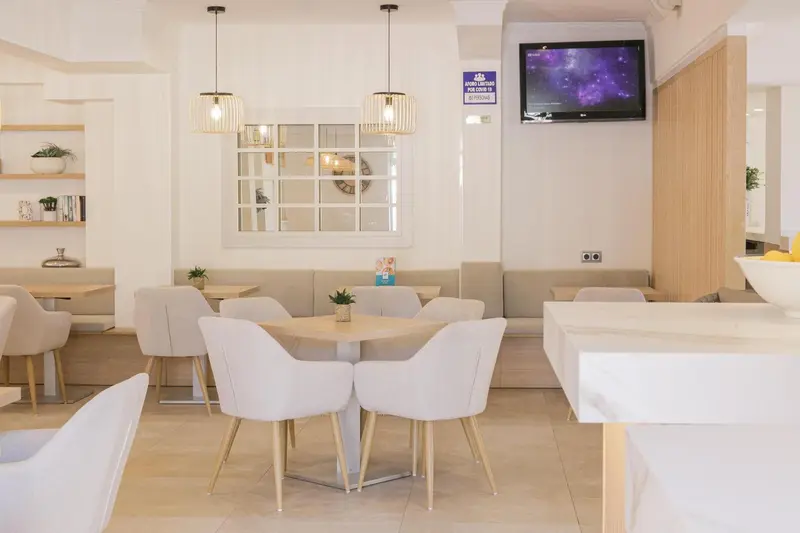 Une salle lumineuse avec des tables et des chaises claires, des suspensions décoratives et un téléviseur mural.