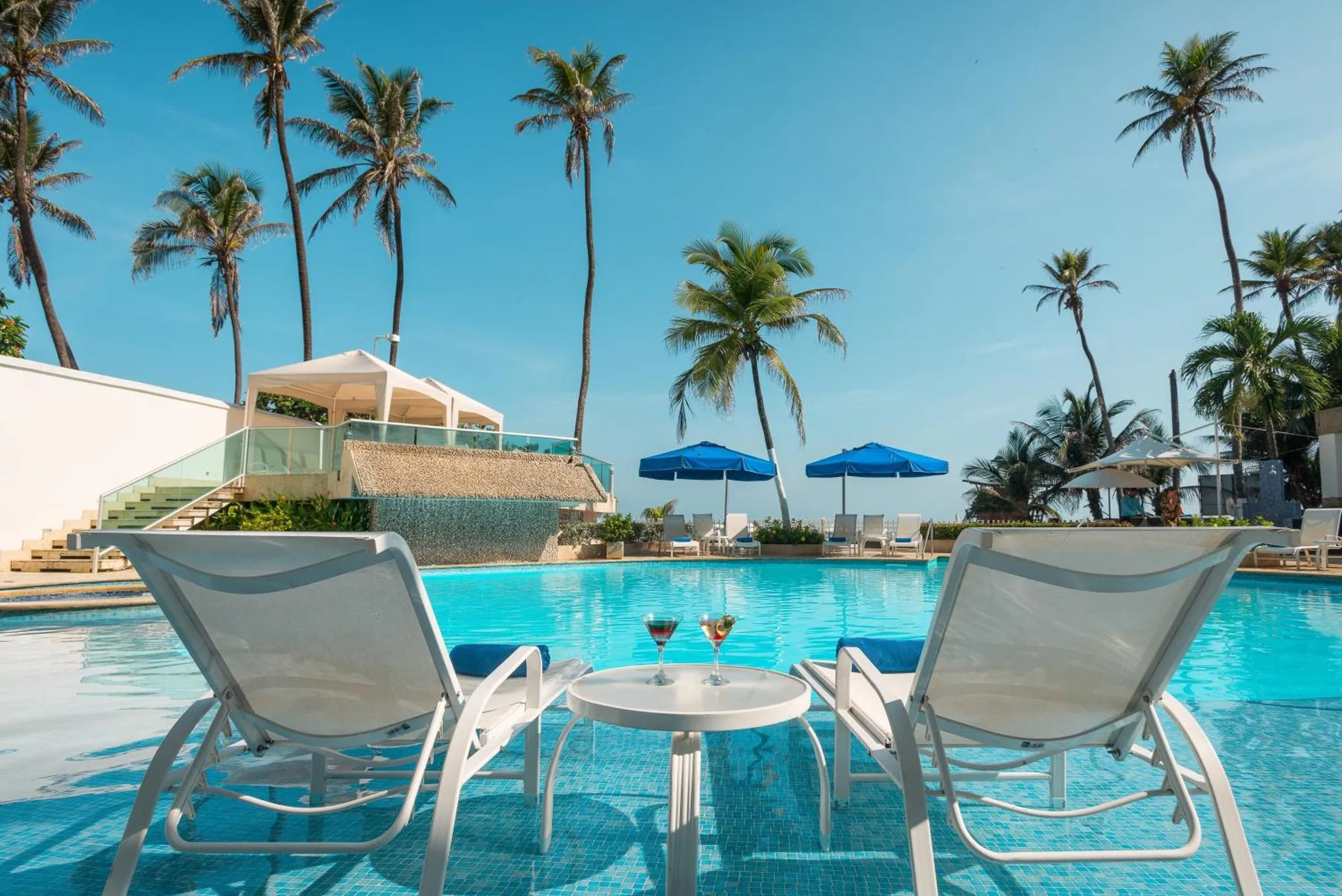 Deux transats blancs sont placés près d'une piscine turquoise, avec des palmiers en arrière-plan sous un ciel bleu.