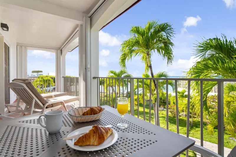 Une table avec un petit-déjeuner est mise en scène sur un balcon avec vue sur la végétation et la mer.