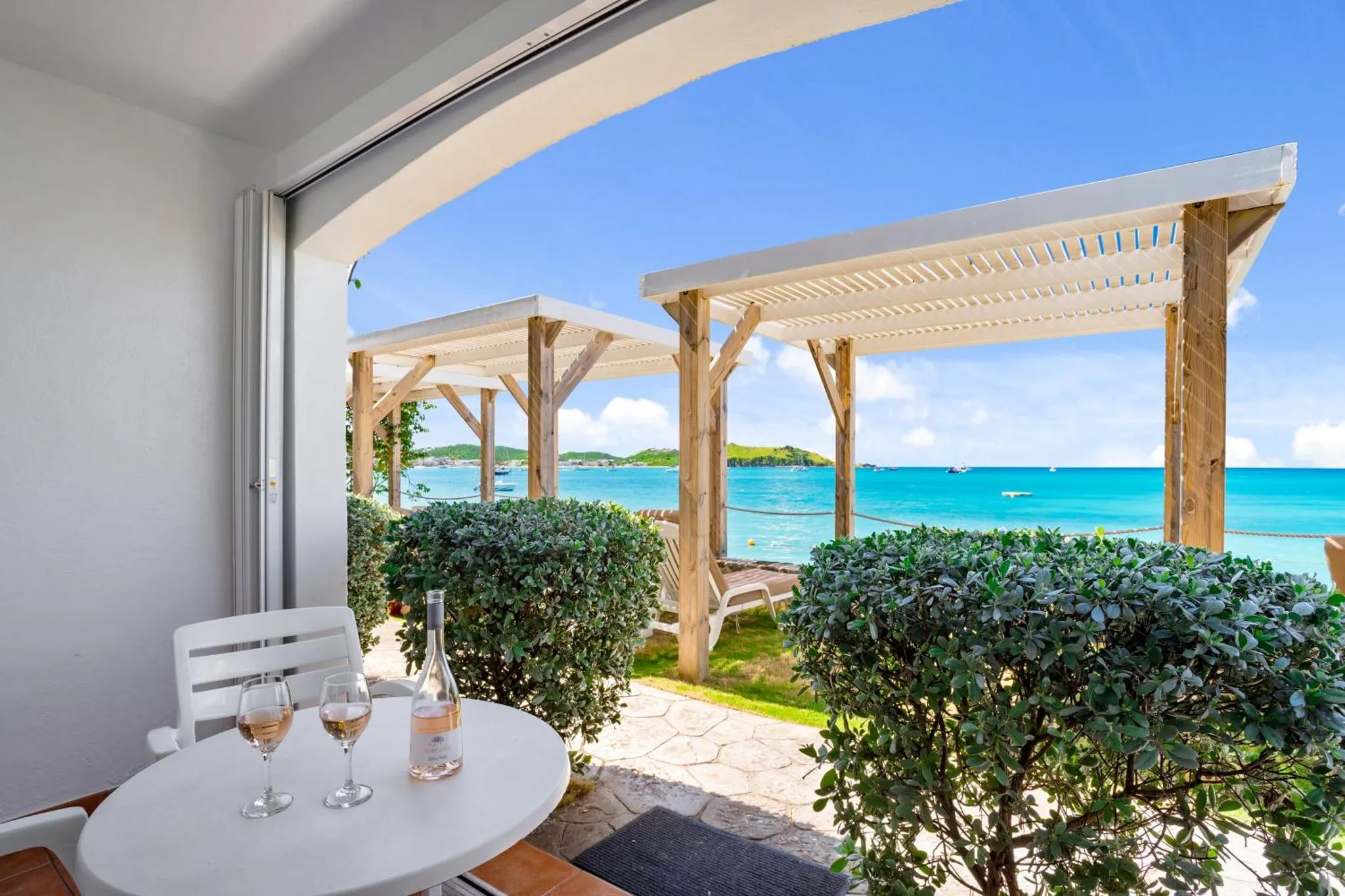 Une table ronde avec des verres de vin est placée devant une terrasse ombragée donnant sur la mer.