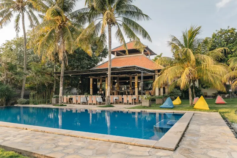 Une piscine rectangulaire bordée d'une terrasse ensoleillée avec des chaises longues et un bâtiment à toit de tuiles en arrière-plan.