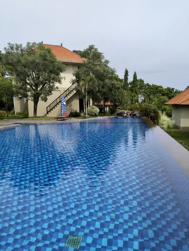 Une piscine bleue rectangulaire avec un bâtiment beige et des arbres en arrière-plan.