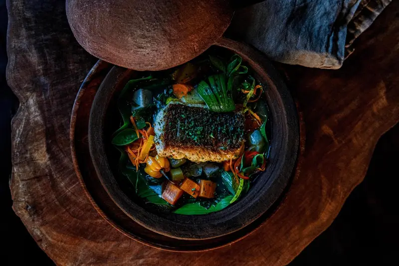 Un plat de poisson cuit est présenté dans un bol en bois rustique, décoré de légumes et d'herbes.