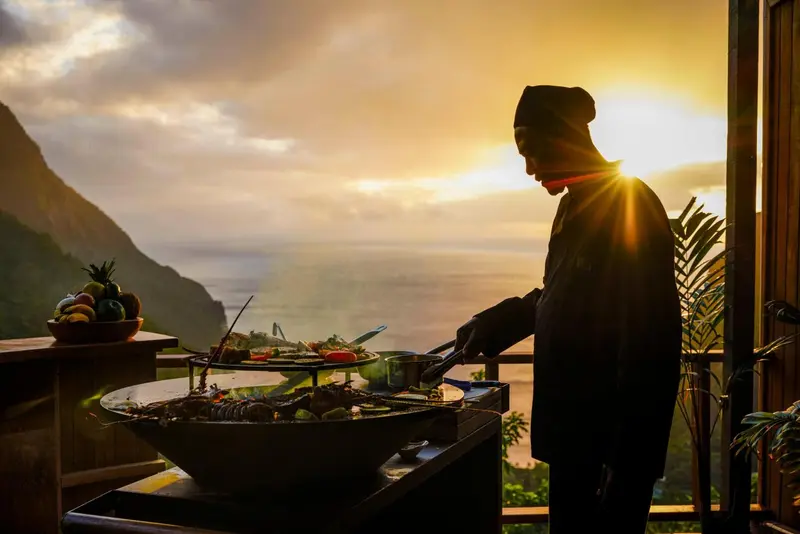 Un chef prépare des grillades sur un balcon avec vue sur la mer et les montagnes.