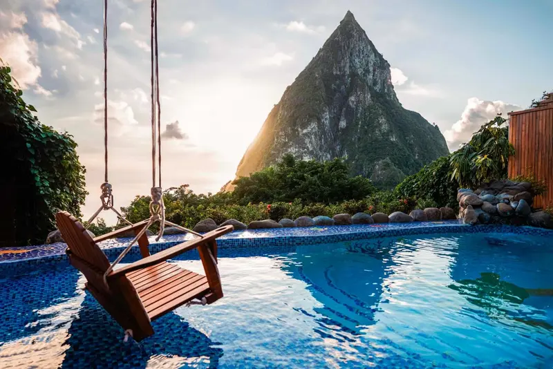 Une balancelle en bois surplombe une piscine turquoise avec une vue sur une montagne.