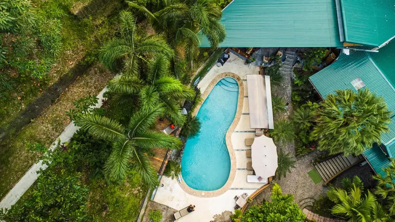 Vue aérienne d'une piscine turquoise entourée de végétation tropicale et de bâtiments.