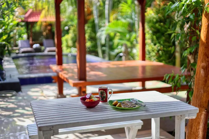 Une table en bois avec un petit-déjeuner est mise en scène devant une piscine et un espace de détente ombragé.