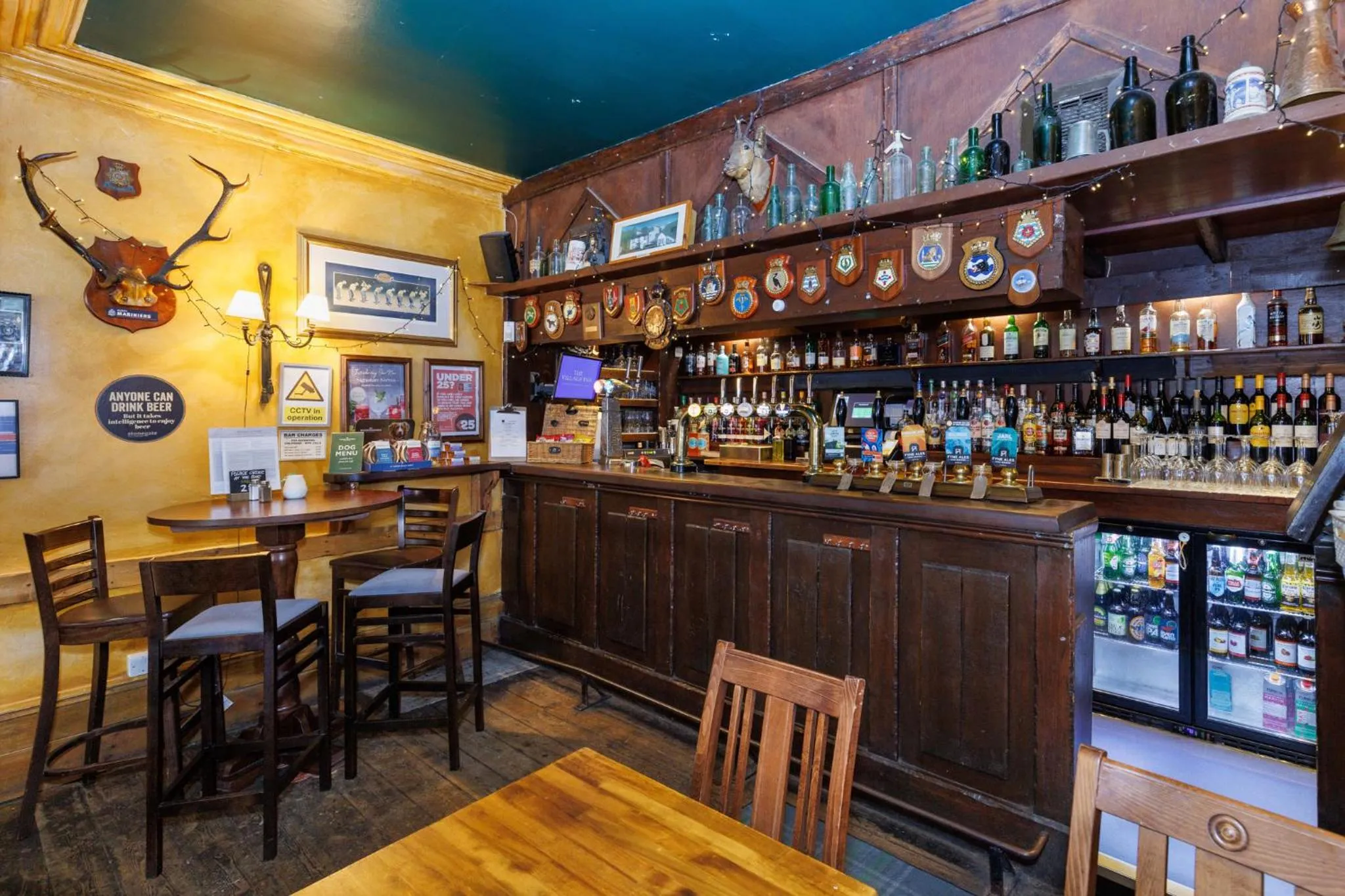 Un comptoir de bar en bois avec des bouteilles et des verres, ainsi que quelques tables et chaises.