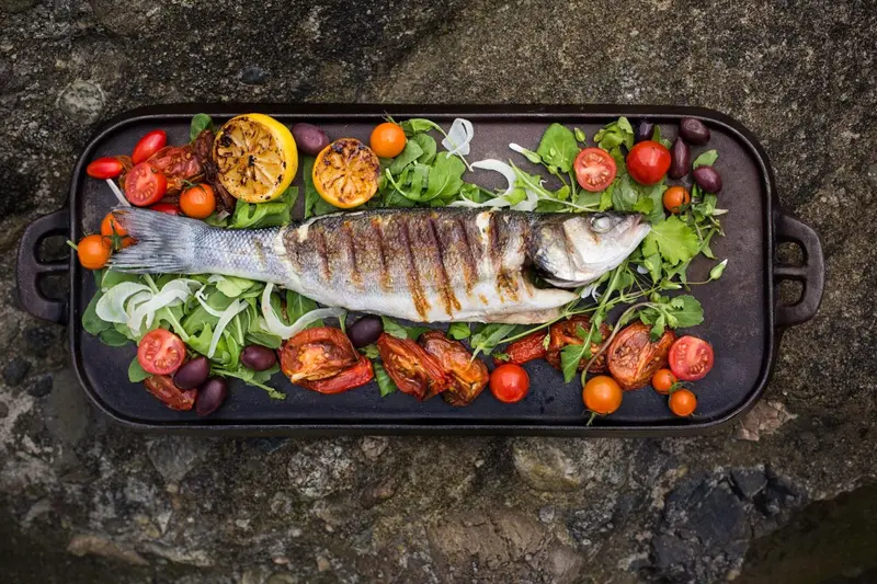 Un poisson grillé est présenté sur un plat rectangulaire noir, décoré de légumes et d'herbes fraîches.