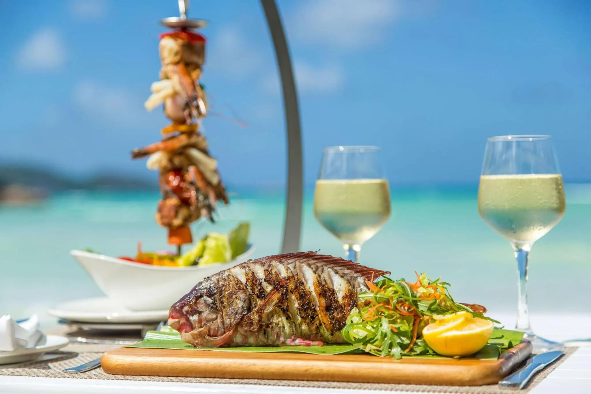 Un plat de poisson grillé est présenté avec une salade et des boissons sur une table en bord de mer.