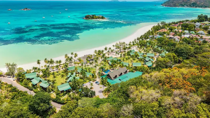 Vue aérienne d'une plage de sable blanc bordée de végétation luxuriante et de bâtiments.