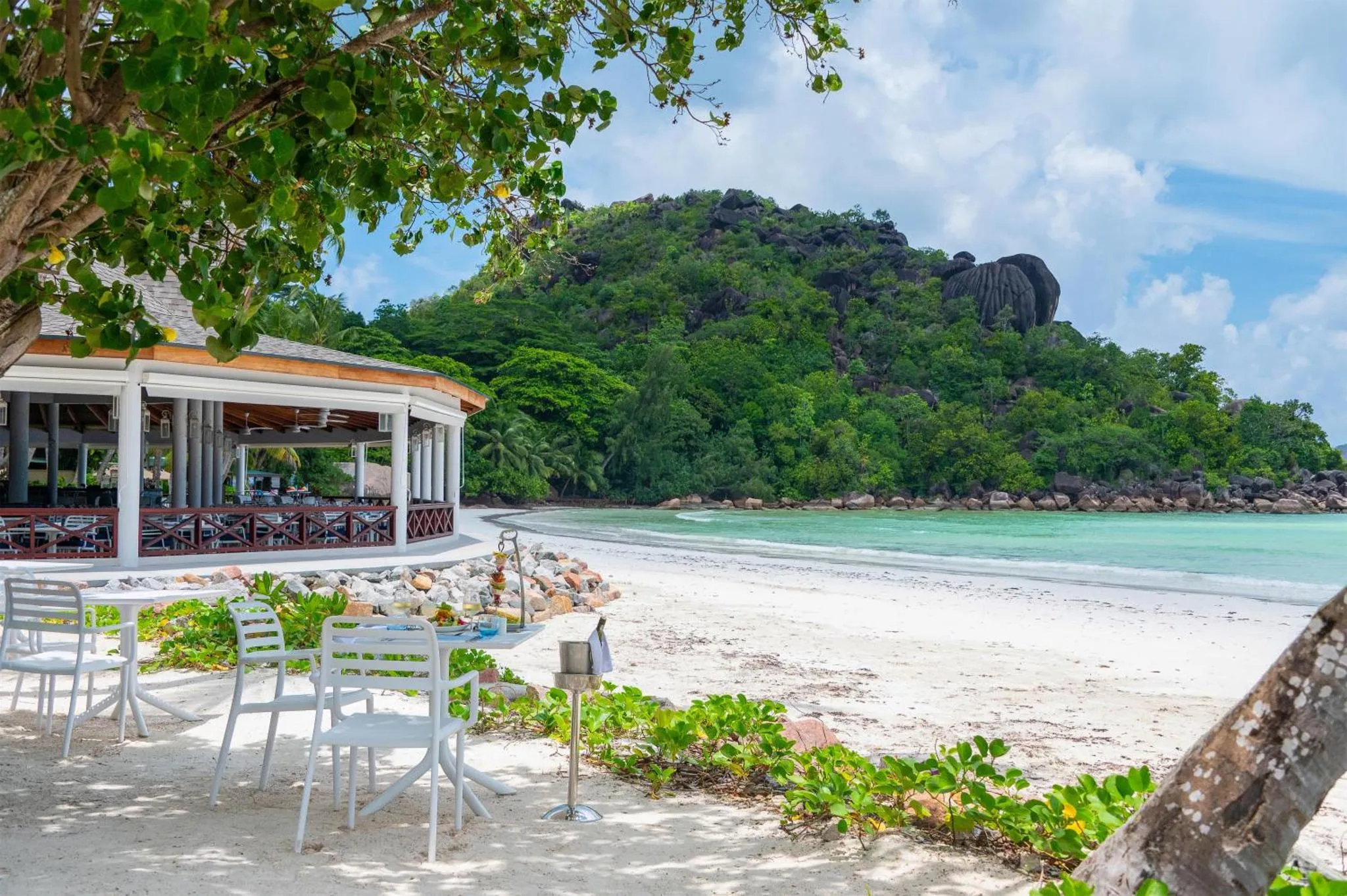 Terrasse de restaurant en bord de mer avec vue sur une plage de sable blanc et une colline verdoyante.