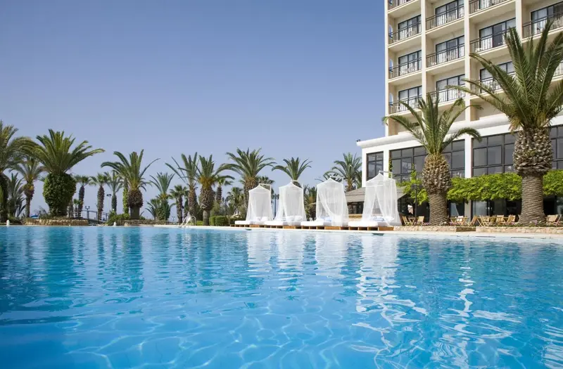 Une piscine turquoise bordée de transats blancs et de palmiers devant un bâtiment beige.