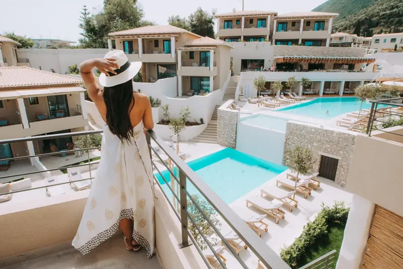 Une femme se tient sur une terrasse surplombant une piscine et des bâtiments blancs.