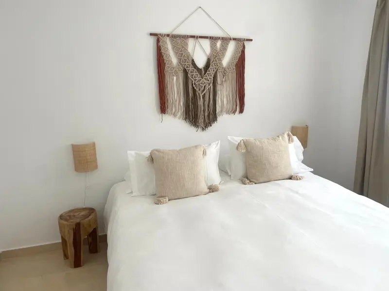 Une chambre lumineuse avec un grand lit recouvert de draps blancs et une décoration murale en macramé.