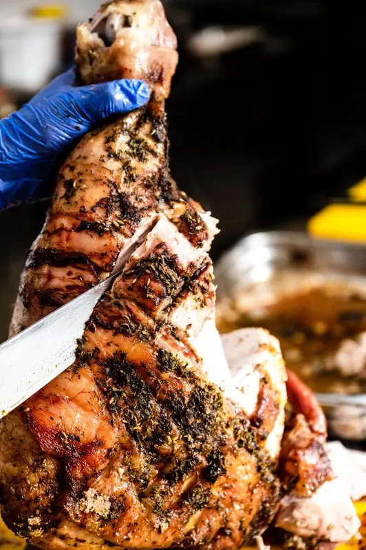 Une main gantée tient une fourchette et un couteau pour dépecer une grosse pièce de viande grillée.